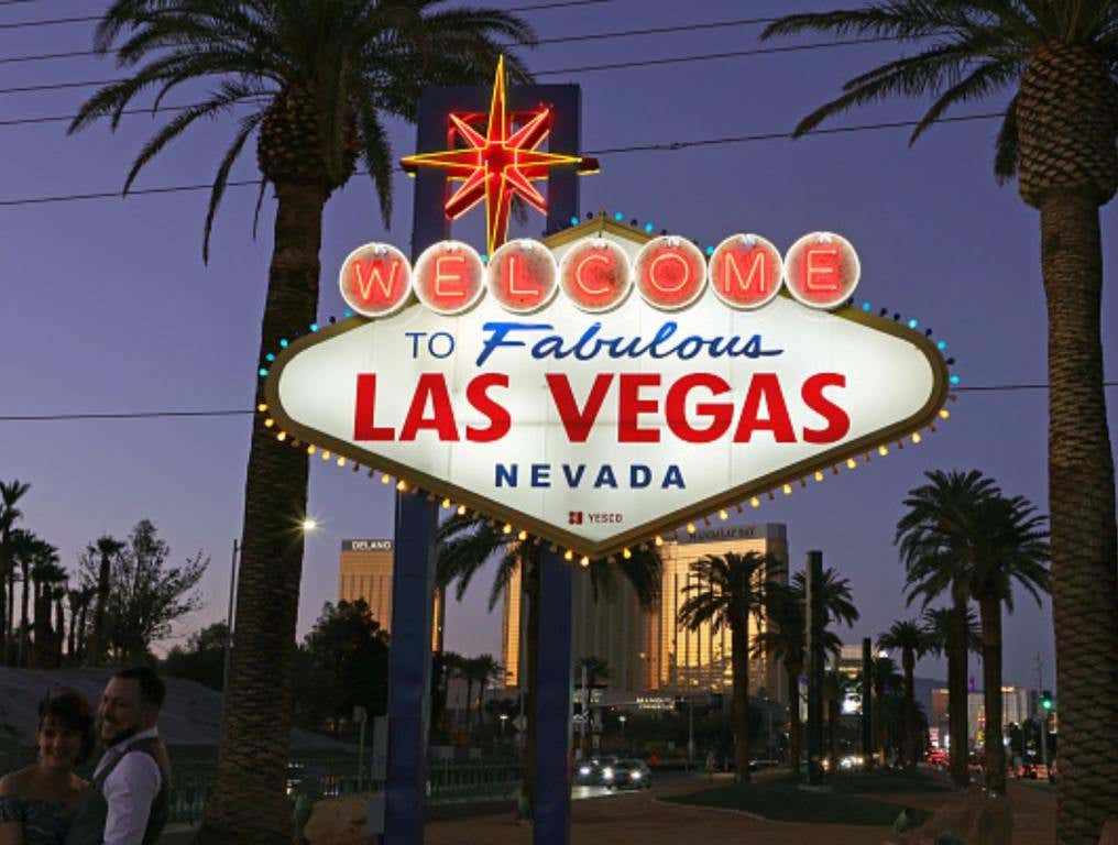 Photo of the iconic "Welcome to Fabulous Las Vegas" sign which welcomes visitors to the Las Vegas Strip. Photo is taken at dusk with the sign lit up. Shadows of nearby palm trees and lit up mega resorts can be seen in the background. Las Vegas is home to four of the most popular casinos in America.