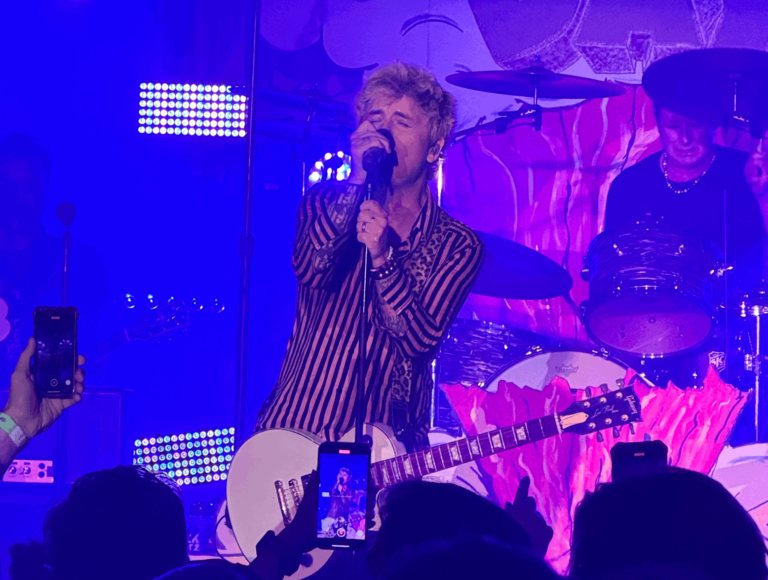 Green Day's Billie Joe Armstrong standing center stage singing into the mic with his eye's closed.