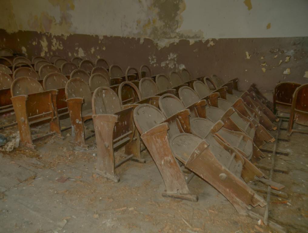 A side angle view of dusty and worn out auditorium seats. Some of the seats have fallen backwards and are leaning against the row behind them. The room they're has a gray concrete floor covered in dust and the gray and brown walls are chipped and damaged.