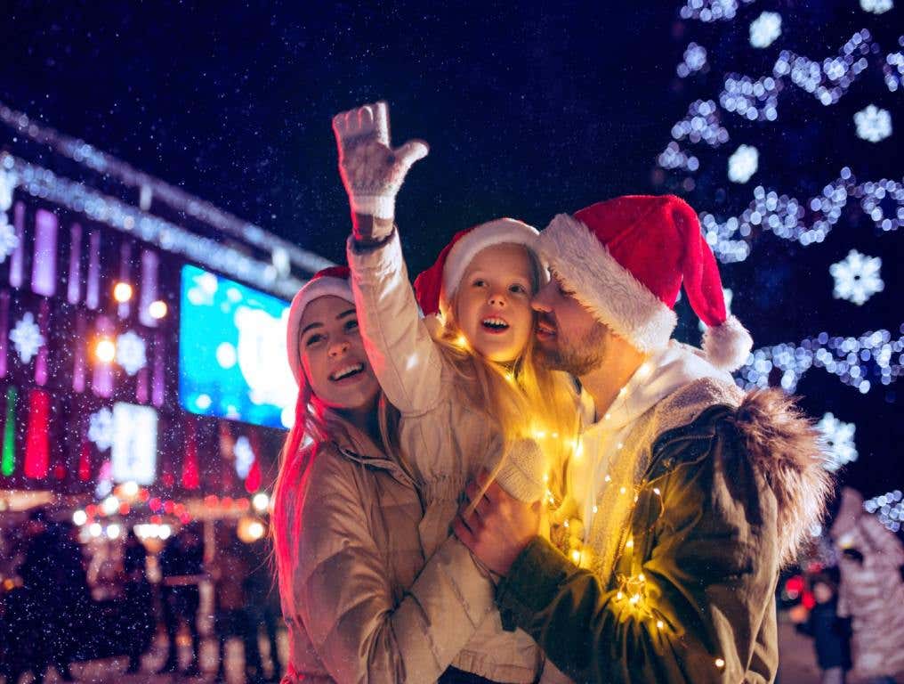 A couple stands in front of a Christmas tree in a strip mall with their young daughter. They are all smiling and wearing Santa hats. Free Vegas Holiday.