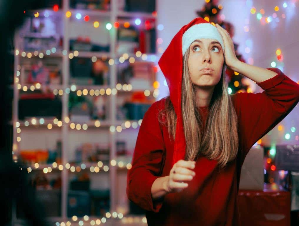 A woman in a Santa hat has her hand on her head and looks up as though she forgot something. She's wearing a red sweater and standing in front of a lit Christmas tree by a window. She needs a last minute Christmas gift.