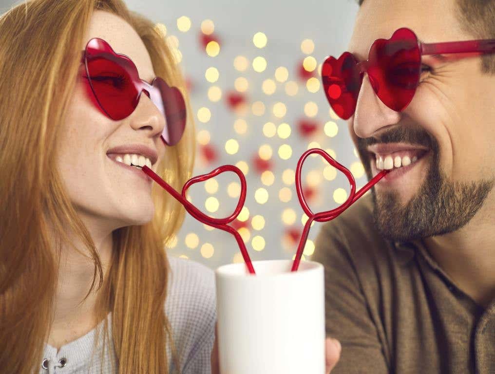 A man and woman wearing heart-shaped glasses look at each other as they share a drink from a single white cup. Both are drinking from heart-shaped straws.