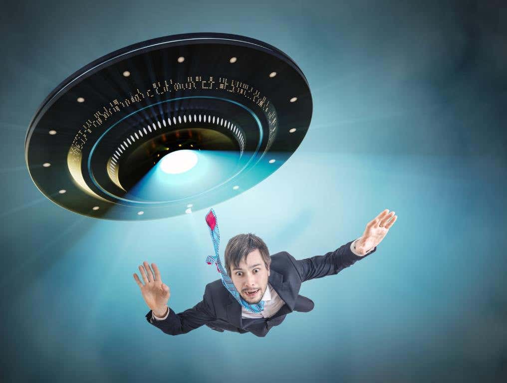 Young man in a business suit and tie floats in midair as he is being pulled in by a UFO tractor beam. Many hope for such an outcome on National Alien Abduction Day.