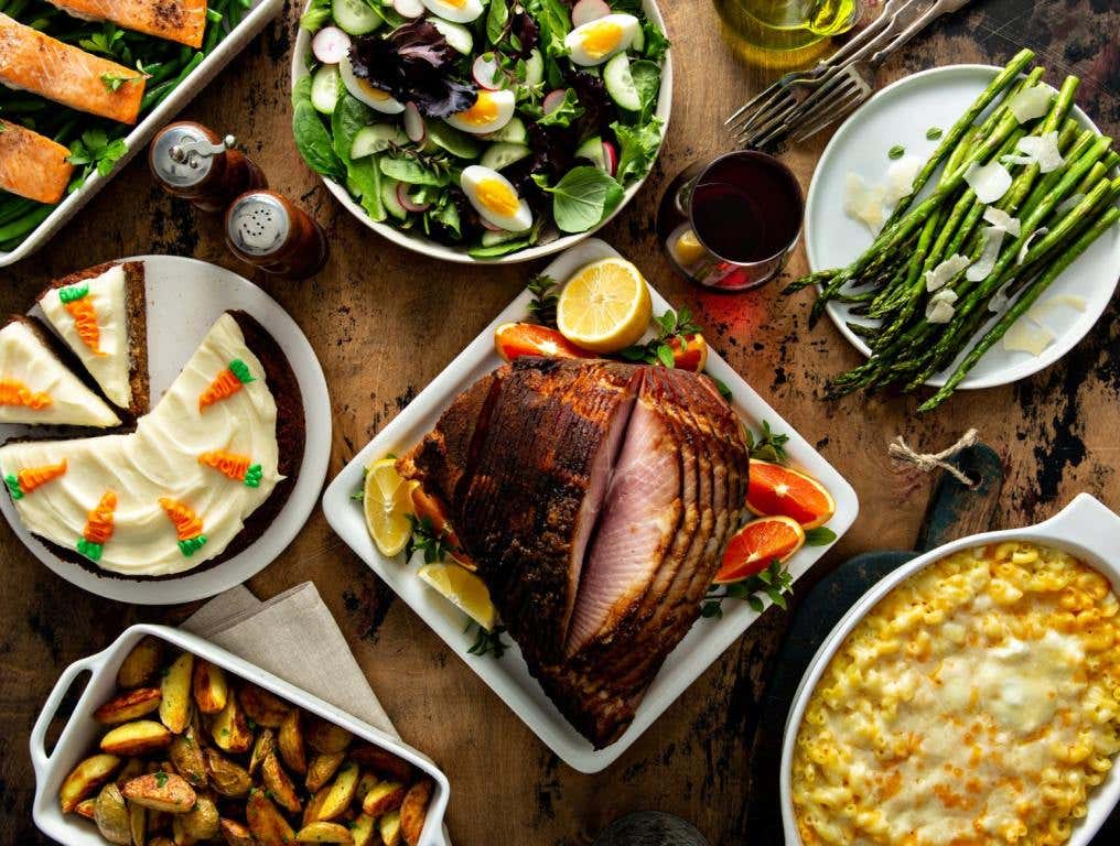 An Easter dinner spread sits on a wooden tabletop. Including a ham in the middle, asparagus, creamed corn, carrot cake and more in dishes surrounding the ham. All dishes are on white plates. Easter in Las Vegas.