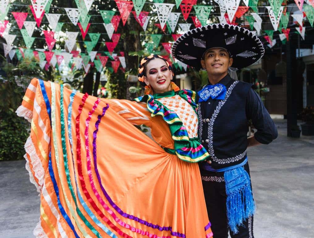 A Hispanic woman (left) is wearing an orange dress with traditional Mexican trim. She is holding her skirt out to the side. On her left is a young Hispanic Man in traditional black Mariachi outfit with a black sombrero. Concept of Cinco de Mayo in Vegas.