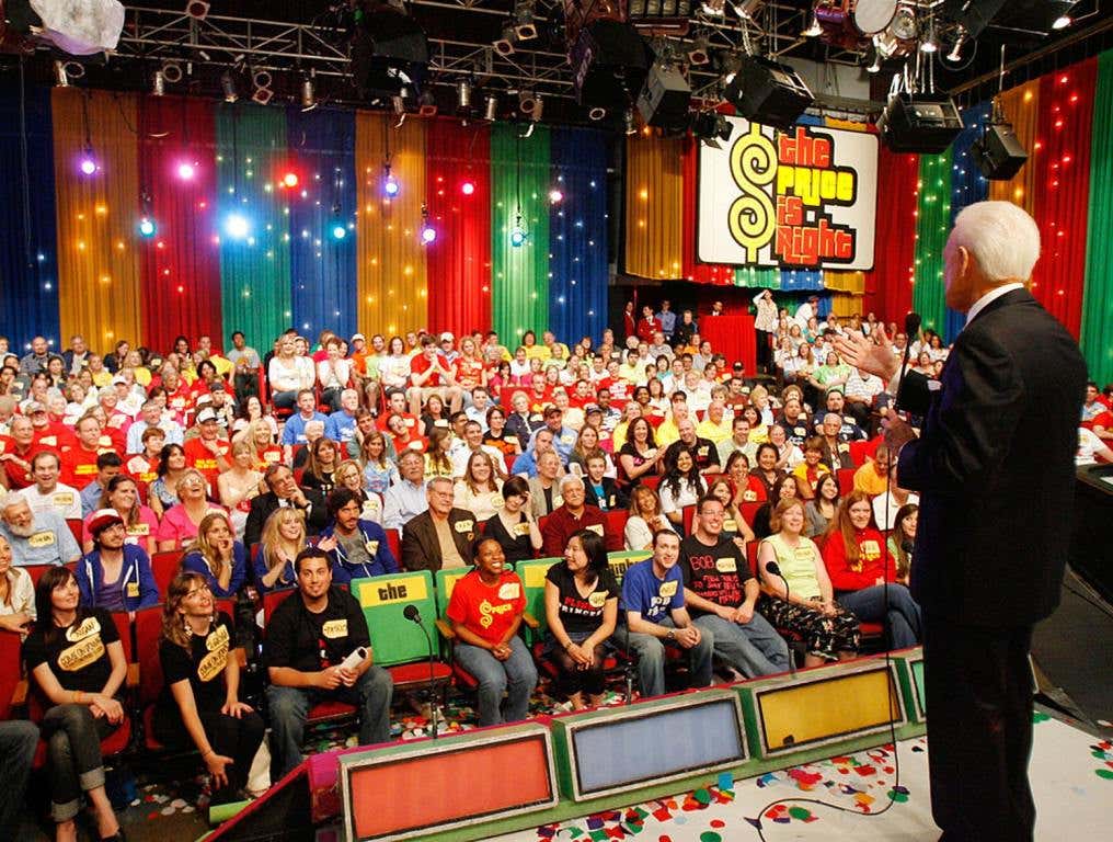 The Price Is Right is coming to Henderson. Photo of legendary host of that TV game show at his final taping in 2007. Bob Barker back and left side are visible as he addresses the audience. He is wearing a dark suit and his hair is white. He is holding a microphone in his left hand and gesturing with his right hand. "The Price Is Right" sign can be seen in the back right side of the studio. The audience is packed with people wearing colorful clothing. The podiums where new contestants stand to make their first bids can be seen at the foot of the stage upon which Bob Barker is standing.