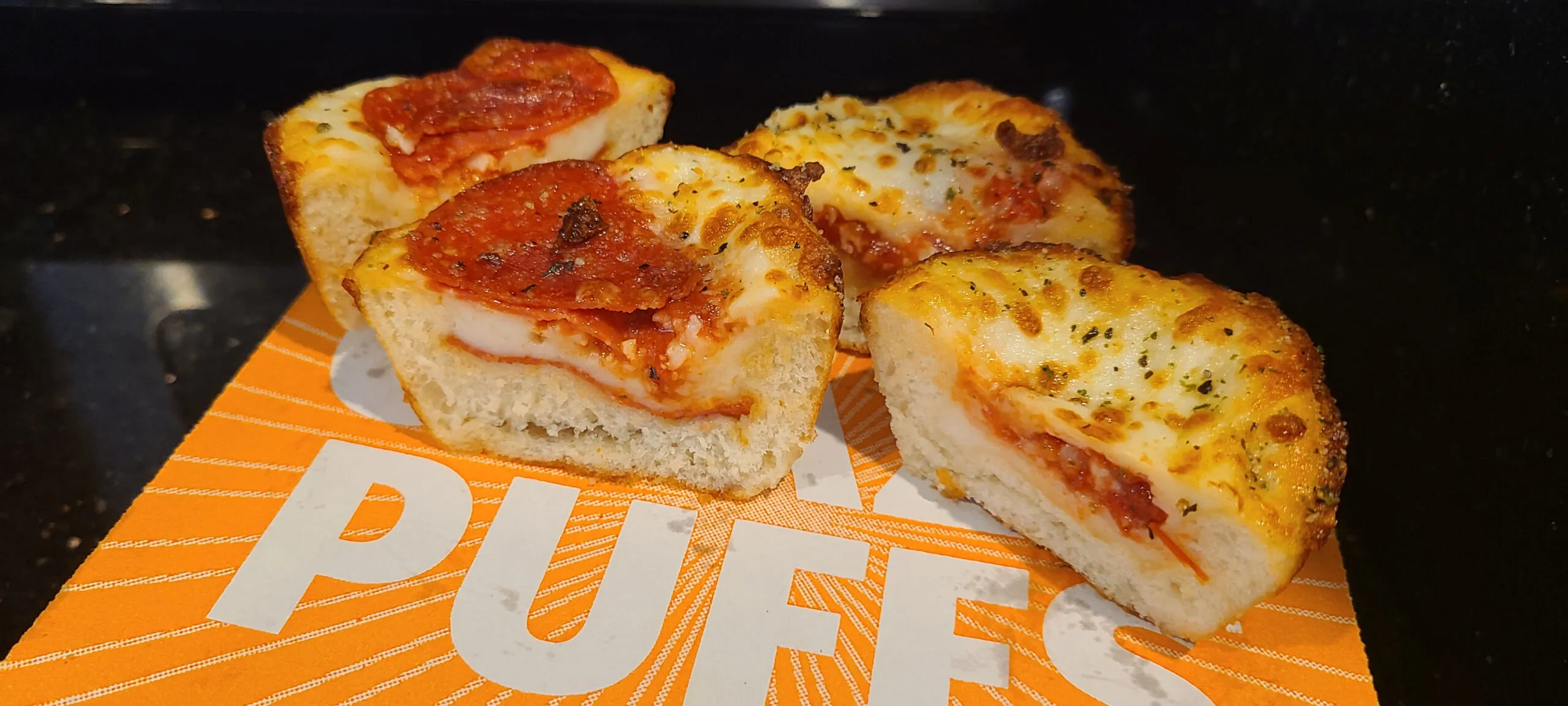 This image shows a Pepperoni Crazy Puff and a Cheese Crazy Puff cut in half and placed on top of a Crazy Puffs box. The puffs have their insides facing the camera to show a cross-section.