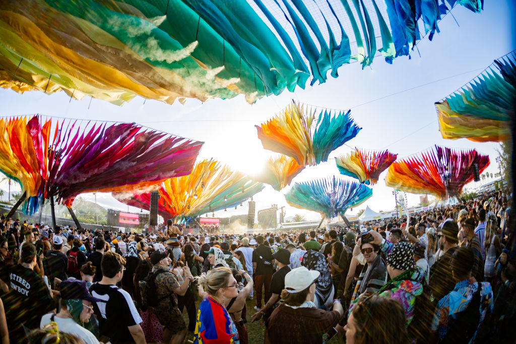 A crowd of people gather under canopies of rainbow-colored fabric. In the background is a stage awaiting a performer for a concert.