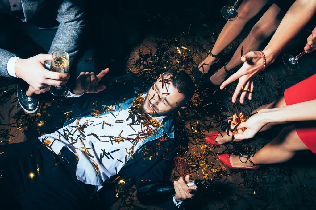 Drunk man in a suit is laying on his back on the floor. He is surrounded by friends with drinks. He is covered in confetti.