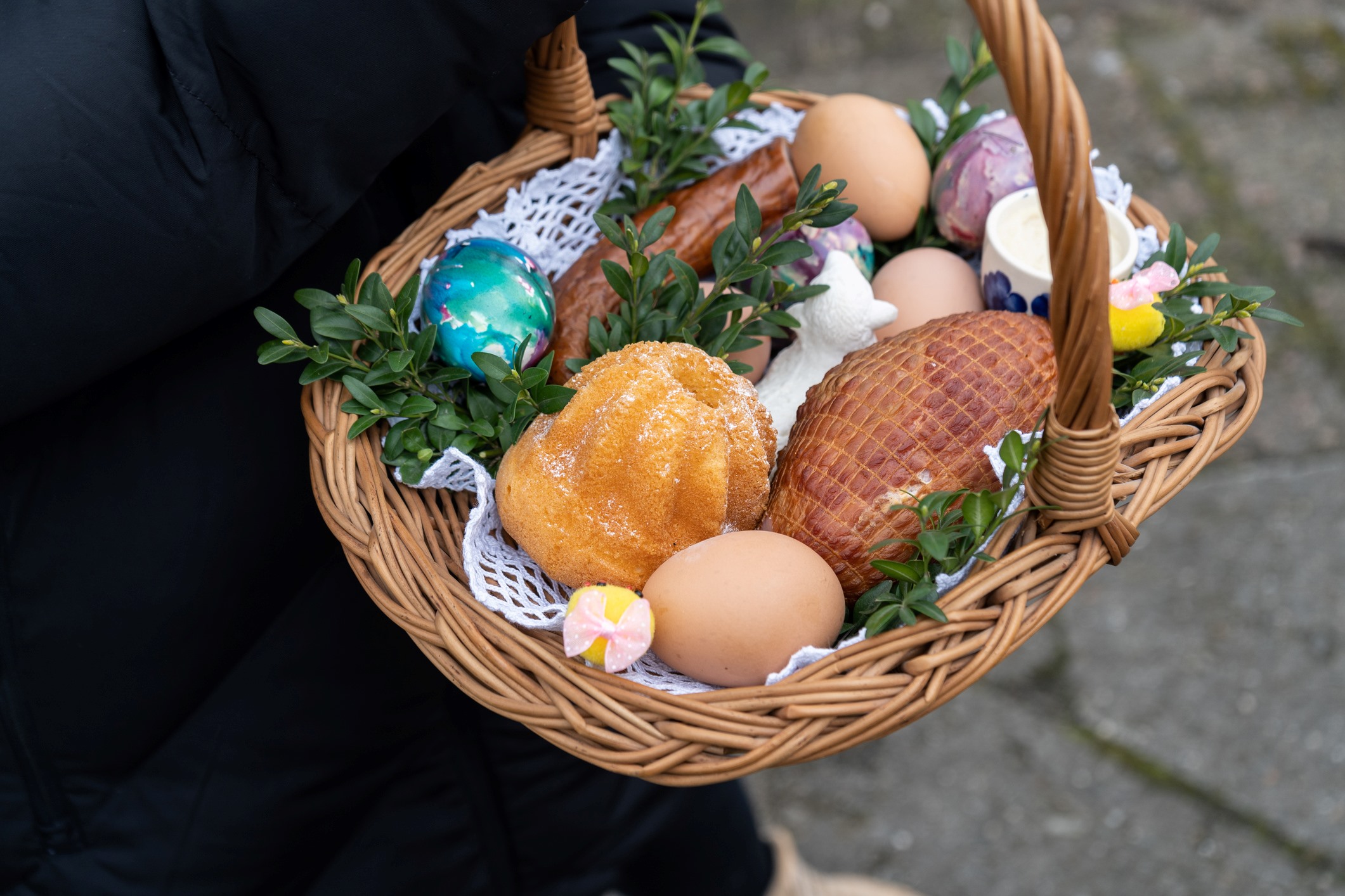 Typical Easter items in an Easter basket. The wicker basket being held by a person dressed in black includes a ham, a sausage, fresh brown eggs, a yellow bundt cake, fragile, decorated glass or plastic easter eggs, a ceramic coffee mug, and decorative green sprigs of fresh herbs.