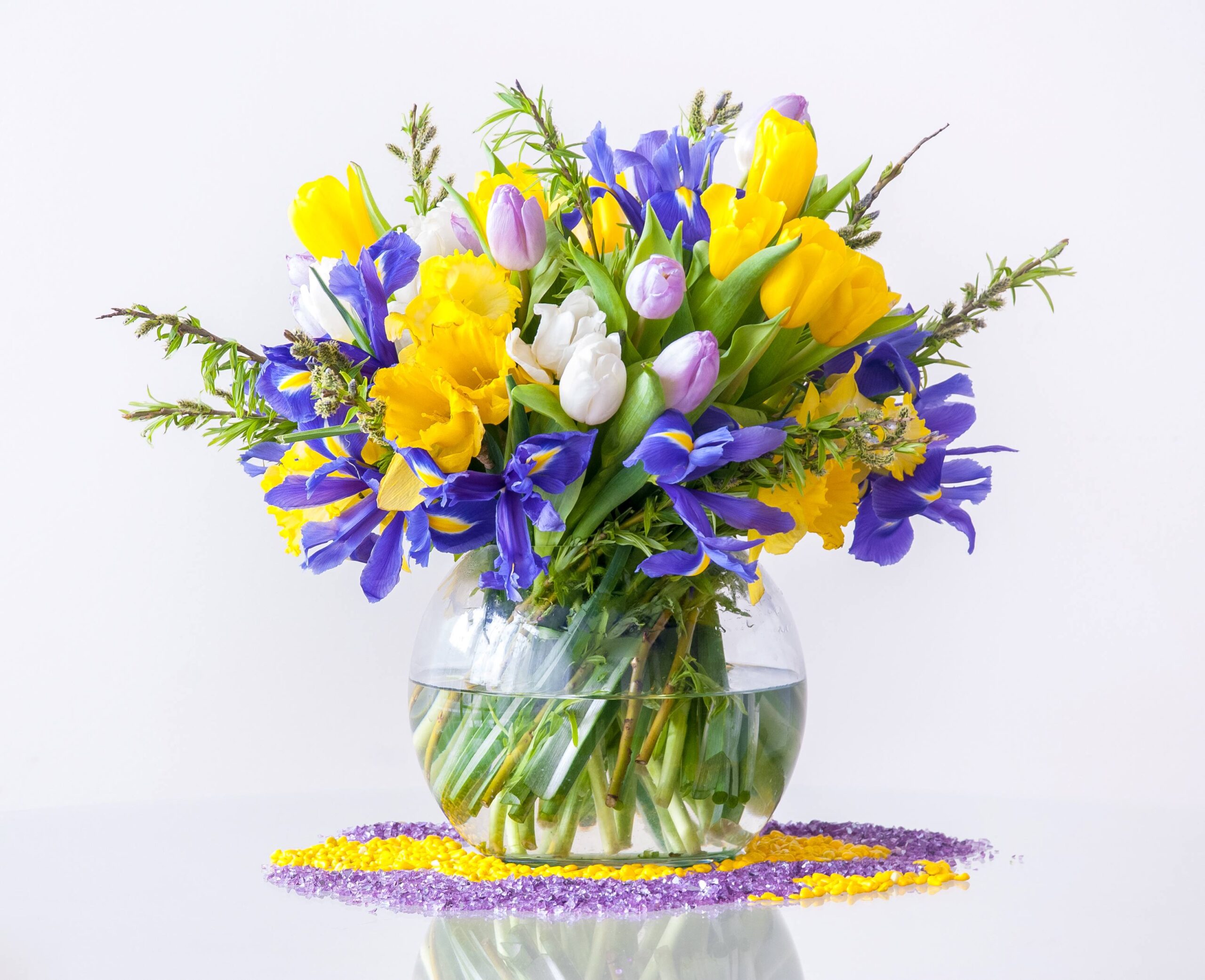 A typical Easter or springtime flower arrangement sitting in a clear, round, squat, bowl, half-filled with water. There is a purple and yellow placemat under the bowl. The cut flowers included in the arrangement are tulips, daffodils, and fleur-de-lises photographed on a white background.