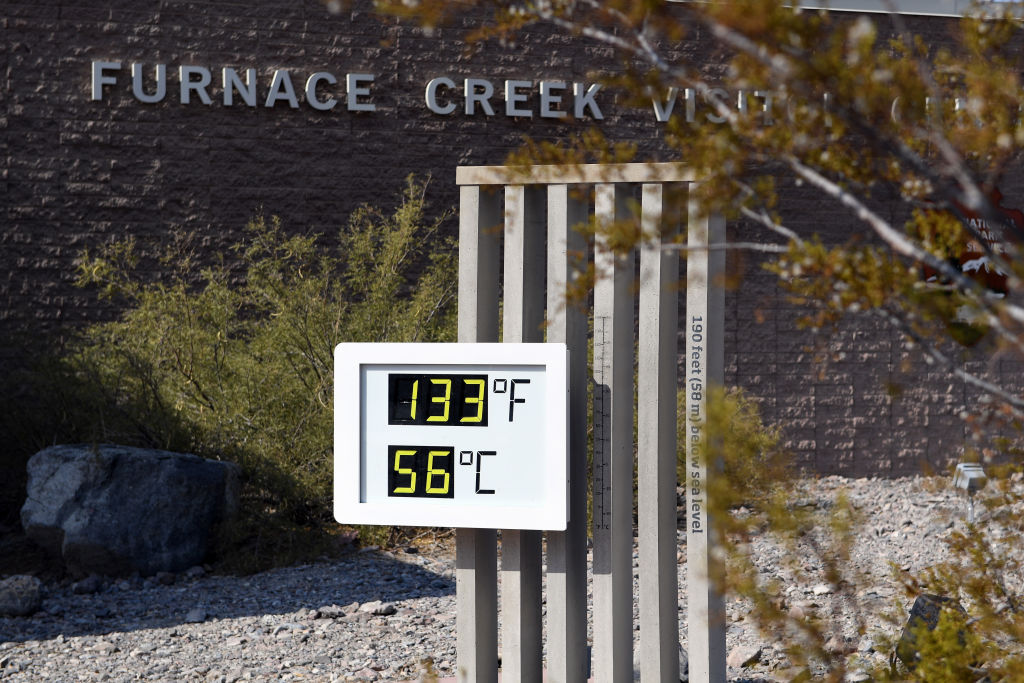 A white board showing digital temperatures hangs on a post. A sign behind it on a brick wall reads "Furnace Creek" in white letters.