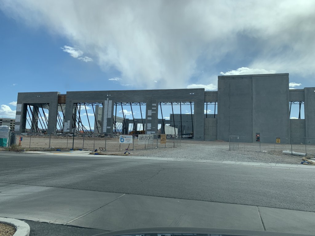 A construction project shows very tall concrete walls with very large openings for windows. The sky above is blue and there are scattered white clouds.