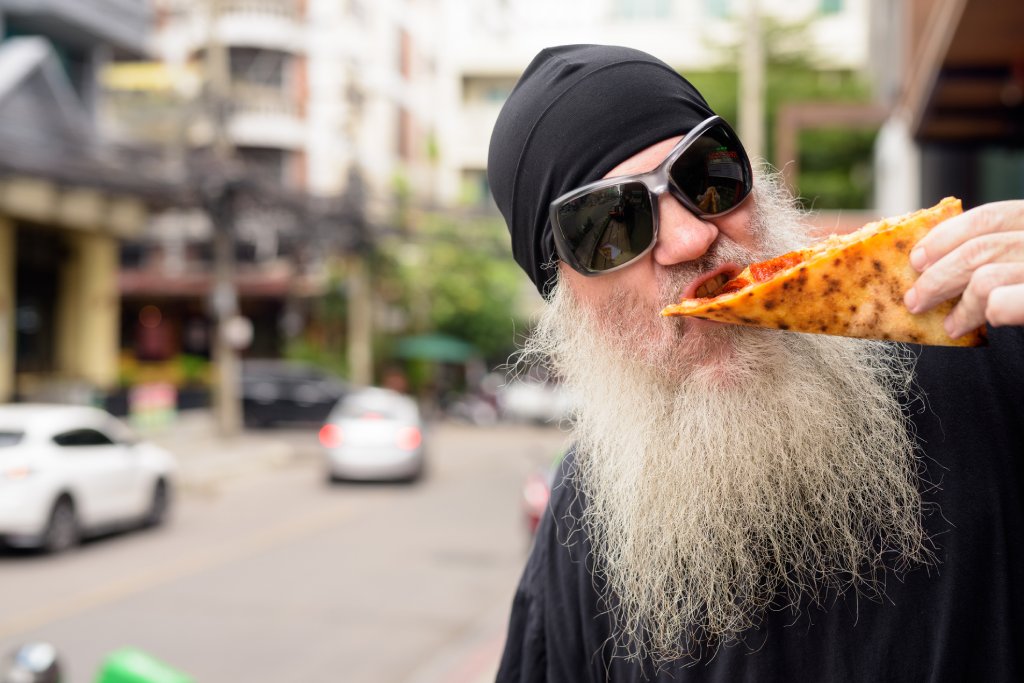 A man in a black beanie and dark sunglasses is eating a folded piece of pizza. He has a black shirt on and long, white beard as he stands outside on a city street.