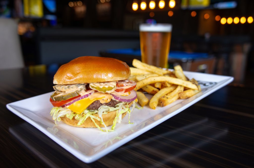 A two-patty burger on a brioche bun with cheese, lettuce, tomato and pickles sits on a white rectangular plate next to a pile of fries. There is a beer in the background behind the plate on the table.