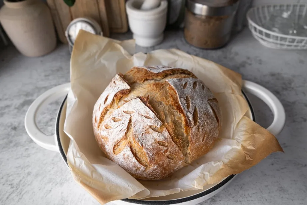 A round loaf of artisan sourdough bread sits in a paper wrapper in a bowl.